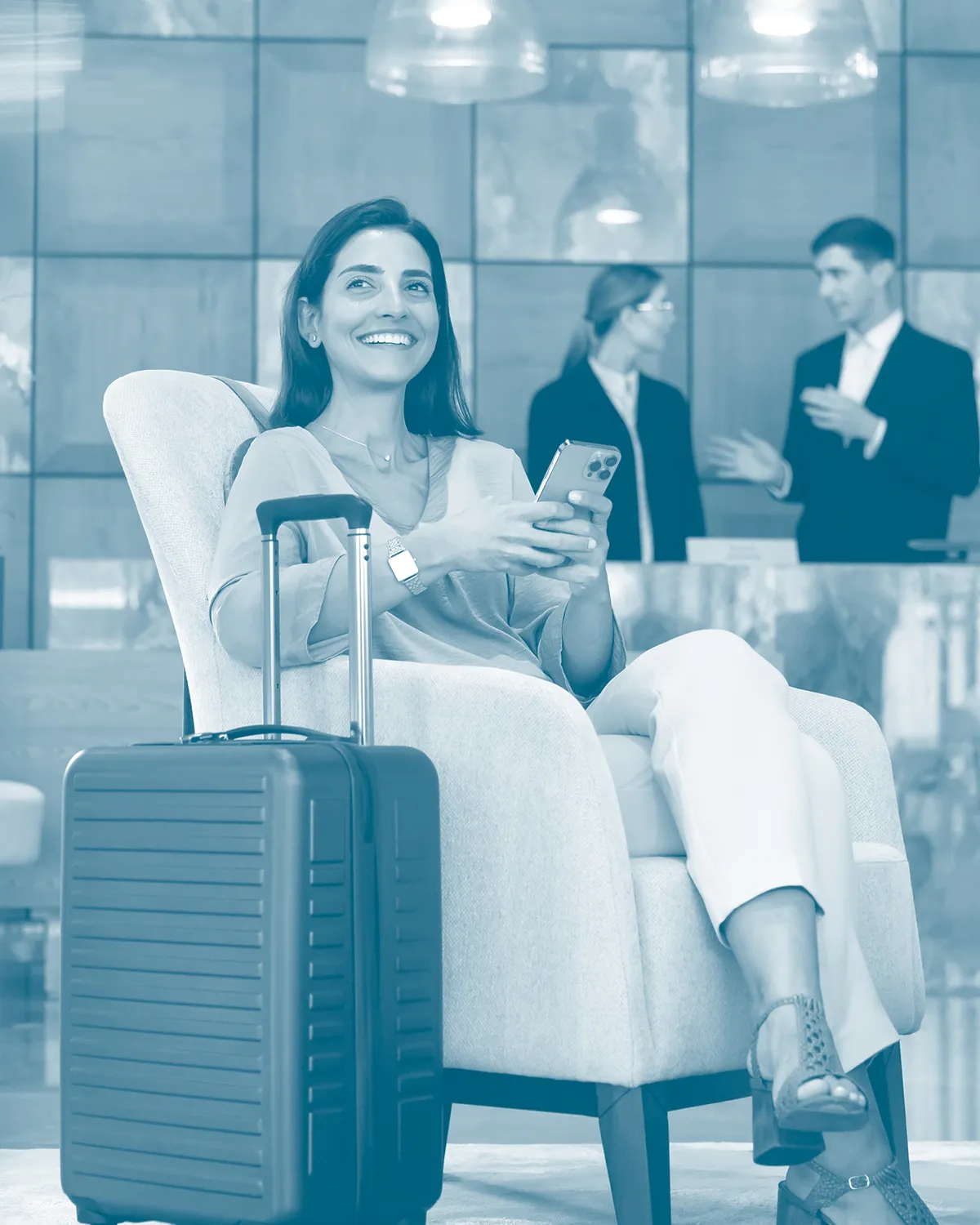A guest seated in a hotel lobby with a suitcase, phone in hand, while staff work at the reception desk behind.