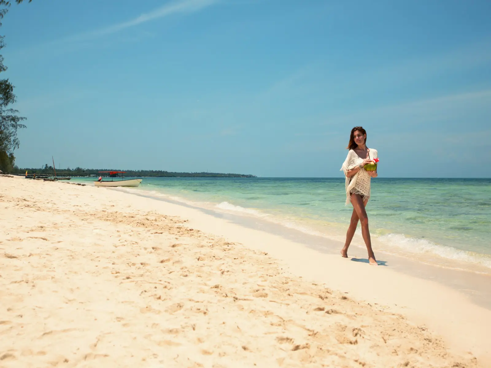 A guest walking the shoreline at Fruit & Spice Wellness Resort with a coconut in hand, a boat moored off the white sand and turquoise water beyond.