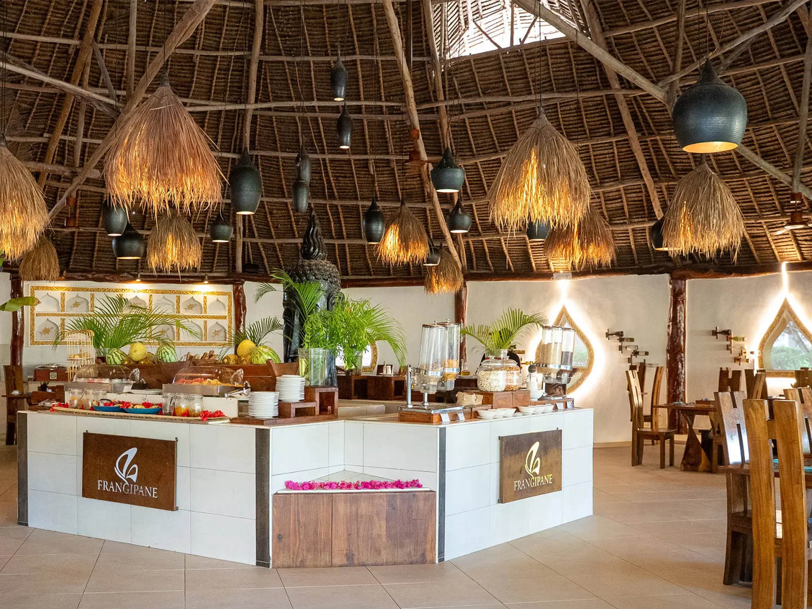 The Frangipane restaurant at Fruit & Spice Wellness Resort: a buffet island laid with fruit beneath a high makuti dome hung with thatch and ceramic pendant lights.