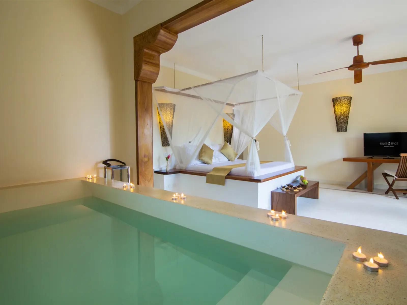 A suite at Fruit & Spice Wellness Resort: a four-poster bed under a white mosquito net, with a candle-lit private plunge pool in the foreground.