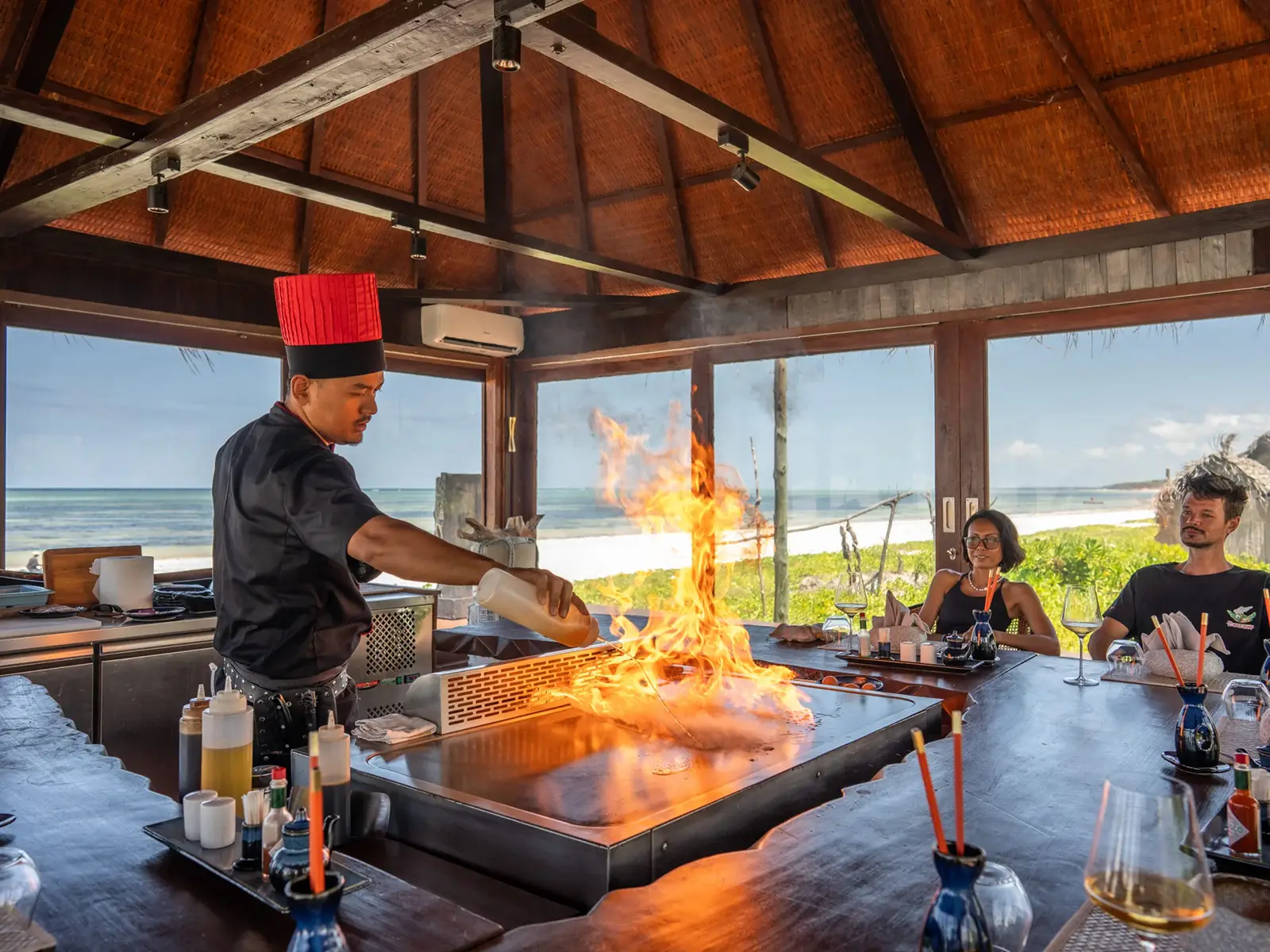 A chef working the teppanyaki grill at Sensations Eco-Chic Hotel, flames rising as guests watch from the counter and the Indian Ocean fills the windows behind.