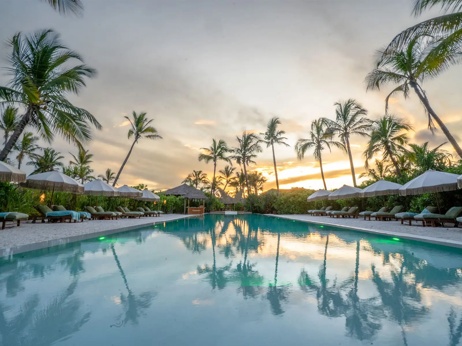 The long pool at Sensations Eco-Chic Hotel at sunset, palms and fringed parasols reflected in the water with loungers down both sides.