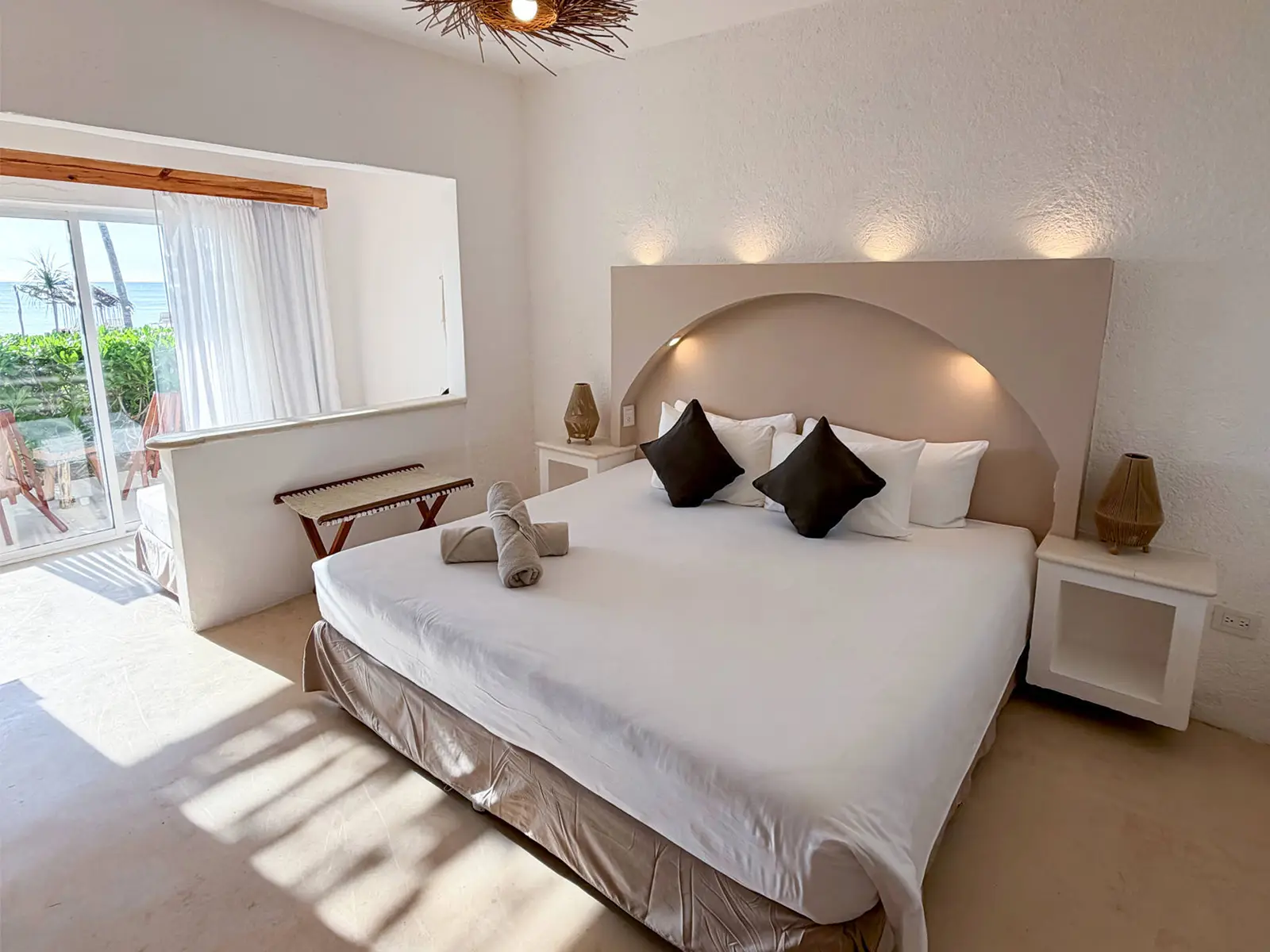 A room at VR Club Tulum Riviera: a king bed against a curved headboard, with a picture window onto the palms and the sea beyond.